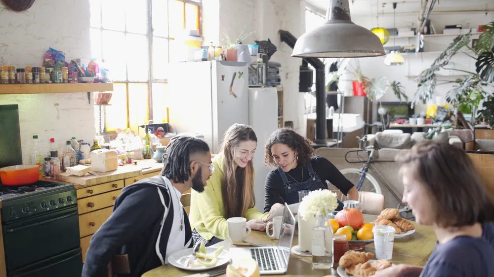 Vier mensen zitten aan een keukentafel met croissants en fruit en kijken samen naar een scherm, in een lichte keuken met veel planten en een groene fornuis
