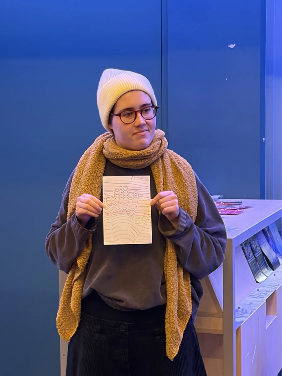 Een persoon met bril, lichte muts en okergele sjaal houdt een vel papier omhoog met daarop een handgetekende optische illusie van een hand, voor een diepblauwe wand