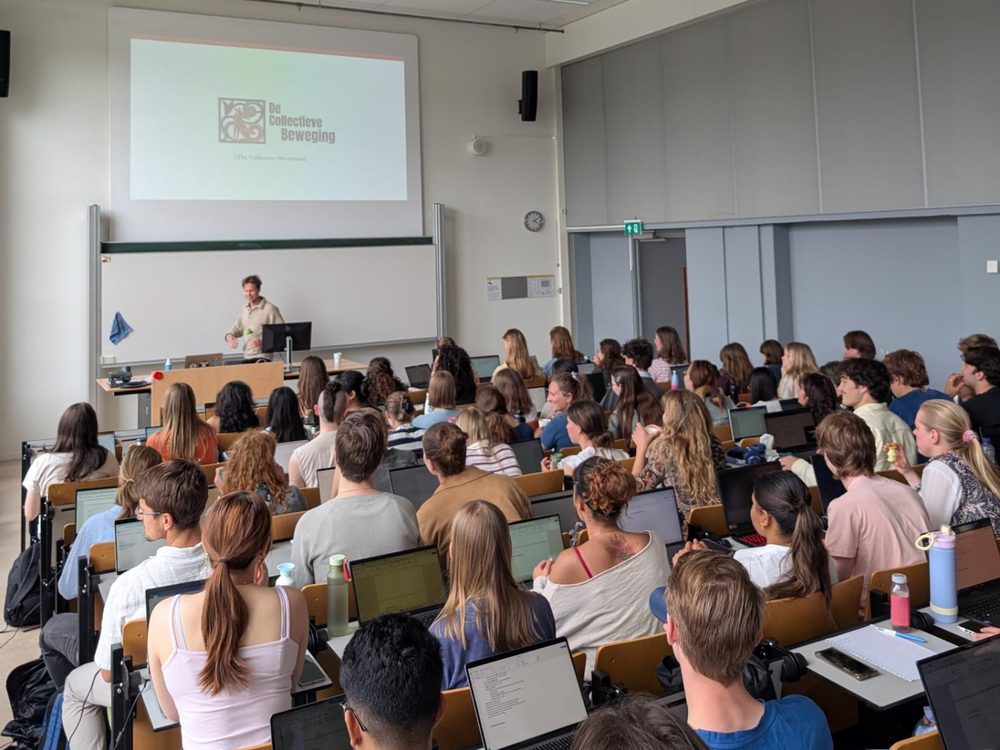 Een spreker geeft een lezing over De Collectieve Beweging in een volle collegezaal